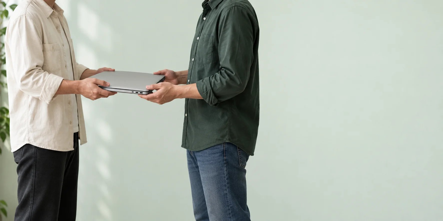 Two people facing each other and handing over a closed silver laptop against a pale green background